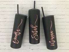 three black tumblers with rose gold writing on them are hanging against a white brick wall