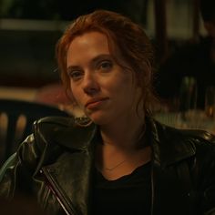 a woman with red hair wearing a black leather jacket looking at the camera while sitting down