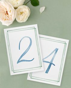 two cards with blue numbers and flowers on green tablecloth next to each other in front of white roses