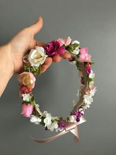 a hand holding a flower wreath with pink, white and purple flowers on it in front of a gray wall