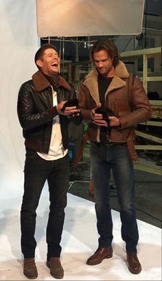 two men standing next to each other in front of a white backdrop and one holding a cell phone