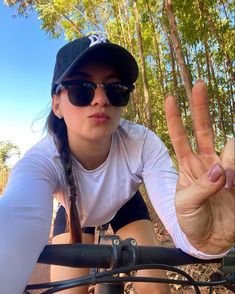 a woman wearing sunglasses and a baseball cap making the peace sign with her hand while riding a bike