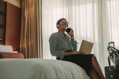 a woman sitting on a bed talking on a cell phone and holding a laptop computer