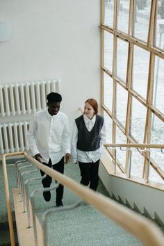 two people are walking down the stairs in front of large windows and looking at each other