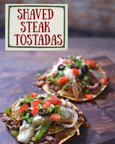 two tortillas topped with meat and vegetables on a cutting board next to a sign that says shaved steak tostadas