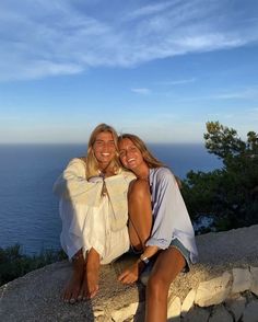 two women are sitting on the edge of a cliff by the ocean and smiling at the camera