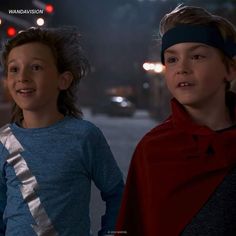 two young boys standing next to each other on a street at night with traffic lights in the background