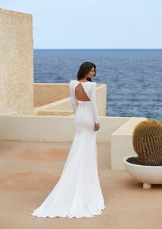 a woman in a long white dress standing next to a cactus and looking at the ocean