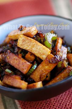 a bowl filled with tofu and vegetables on top of a red table cloth next to a wooden spoon