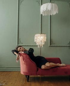 a woman laying on top of a red couch under a chandelier next to a lamp