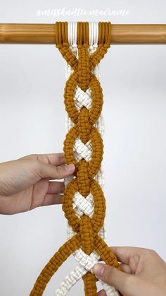 two hands are working on an orange and white crochet piece that is hanging from a wooden pole