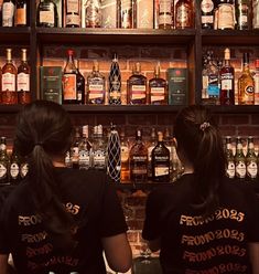 two women in black shirts behind a bar with liquor bottles on the shelves above them