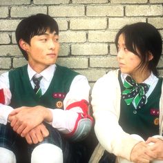 two people sitting next to each other in front of a brick wall wearing vests and ties