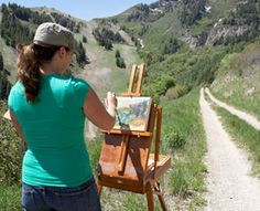 a woman is painting on an easel in the mountains