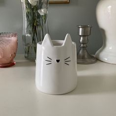 a white cat mug sitting on top of a table next to a vase with flowers in it