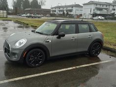 a grey mini cooper parked in a parking lot on a rainy day with houses in the background