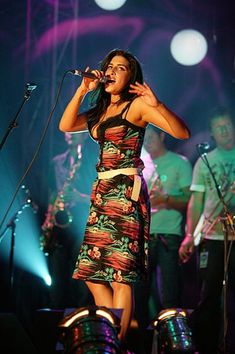 a woman singing into a microphone on stage with other people in the background at a concert