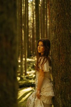 a woman in a white dress standing next to a large tree with moss growing on it