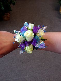 a wrist corsage with white roses, purple and green flowers on it is being held by someone's arm