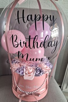 a pink birthday cake with balloons and flowers in a glass box on a chair that says happy birthday mum