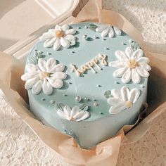 there is a blue cake with white flowers on it in a cardboard box next to lace doily