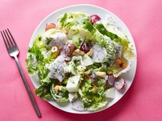 a white plate topped with a salad next to a fork and knife on a pink surface