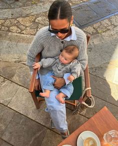 a woman holding a baby while sitting in a chair next to a cup of coffee