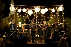 a group of people standing on top of a stage with lights strung over them and playing instruments