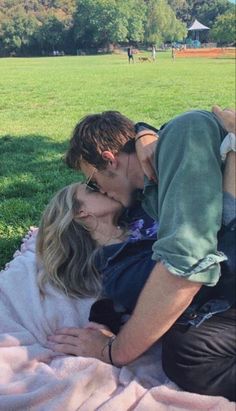 a man and woman kissing while laying on a blanket in the grass at a park