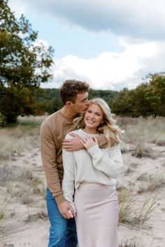 a man and woman standing next to each other in the sand with trees behind them