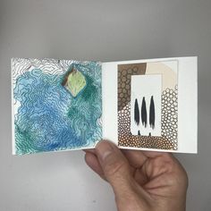 a hand is holding an open book with drawings on the pages and watercolors