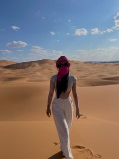 a woman in white pants and pink bandana walking through the desert with her back to the camera