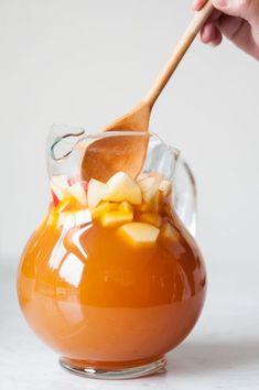 a person holding a wooden spoon in a glass pitcher filled with liquid and apples on the side