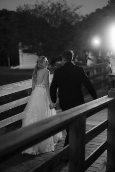 a bride and groom walking down a wooden bridge at night with lights on the other side