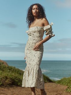 a woman in a white dress standing on a dirt road near the ocean with her hand on her hip