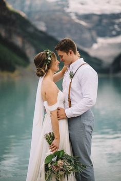 a bride and groom standing next to each other in front of a lake with snow covered mountains