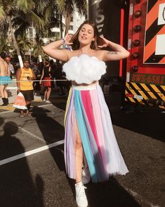 a woman in a rainbow colored skirt and crop top is standing on the street with her hands behind her head