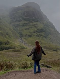 a woman standing on top of a lush green hillside next to a tall mountainside