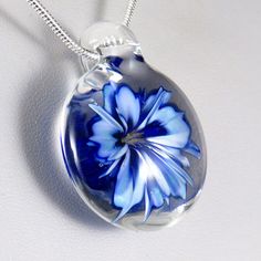 This unusual and unique pendant has a realistic blue and white glass 3D flower inside a solid glass - this isn't resin or a real flower, it's all made entirely of glass! I've used a cobalt blue over white, which I've graduated in tone and to form bands on the petals to make the flower look more realistic, also giving the edges and centre a darker rim of blue. In the centre I've added some yellow 'stamens' to make it look more realistic, where some bubbles have clustered around them to add a dew White Flower Shaped Glass Jewelry, Blue Glass Flower Shaped Jewelry, Blue Flower Shaped Glass Jewelry, Blue Flower-shaped Glass Jewelry, Silver Glass Flower Pendant Jewelry, Blue Round Pendant Jewelry With Flower Charm, Blue Flower Pendant Necklace For Gift, Blue Flower Charm Round Pendant Jewelry, Blue Flower Pendant Necklace With Large Pendant