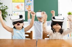 two children wearing virtual headsets while sitting at a table with their hands in the air