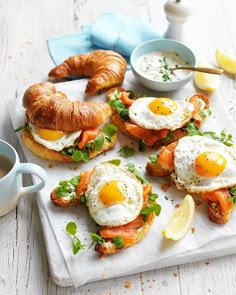 breakfast sandwiches with eggs, salmon and croissants on a white tray next to a cup of coffee