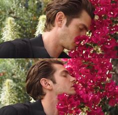 a man with his eyes closed standing in front of red flowers and smelling the flower