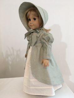 a doll dressed in a dress and bonnet is standing on a white table with a white wall behind it