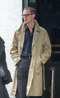 a man wearing glasses and a trench coat is standing in front of a door with his hands in his pockets