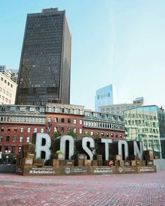 the word boston spelled out in front of some tall buildings and bricked pavement with people walking around it