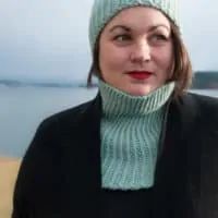 a woman wearing a knitted hat and black coat by the water with her eyes closed