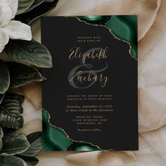 a black and gold wedding card with greenery on it, surrounded by white flowers