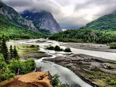 a river flowing through a lush green forest filled valley next to mountains covered in clouds