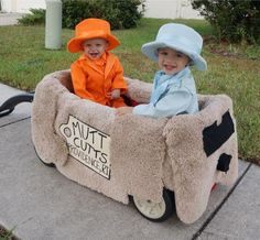 two young children sitting in a fake cow cart on the sidewalk with grass and bushes behind them