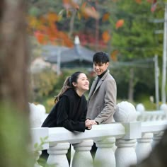 a man and woman standing next to each other on a white fence with trees in the background